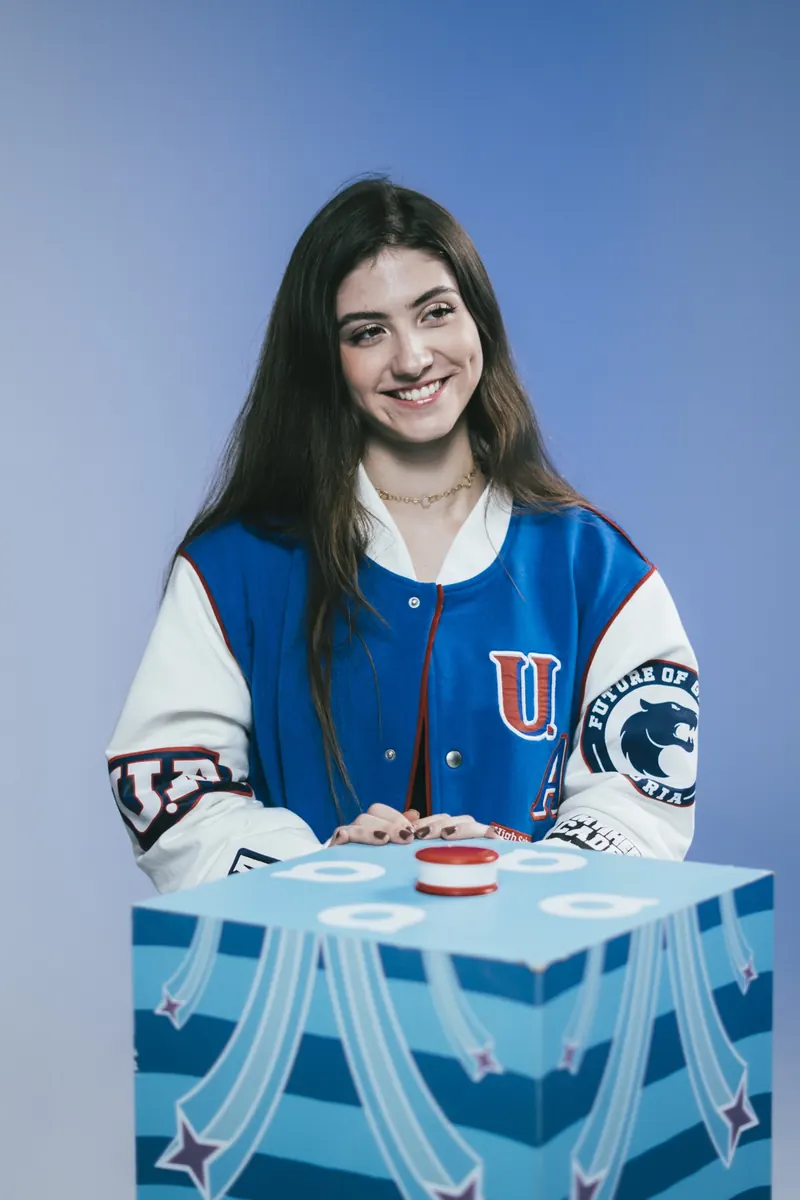 Mulher sorrindo, de jaqueta azul e branca, atrás de um display em forma de cubo azul com um botão vermelho em cima.
