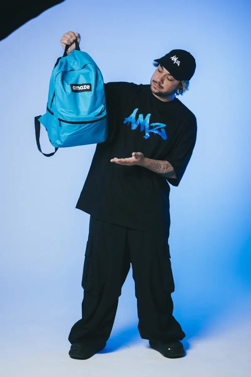 Homem de boné e camiseta preta segura no alto uma mochila azul-turquesa e aponta para ela com a outra mão, em fundo azul.