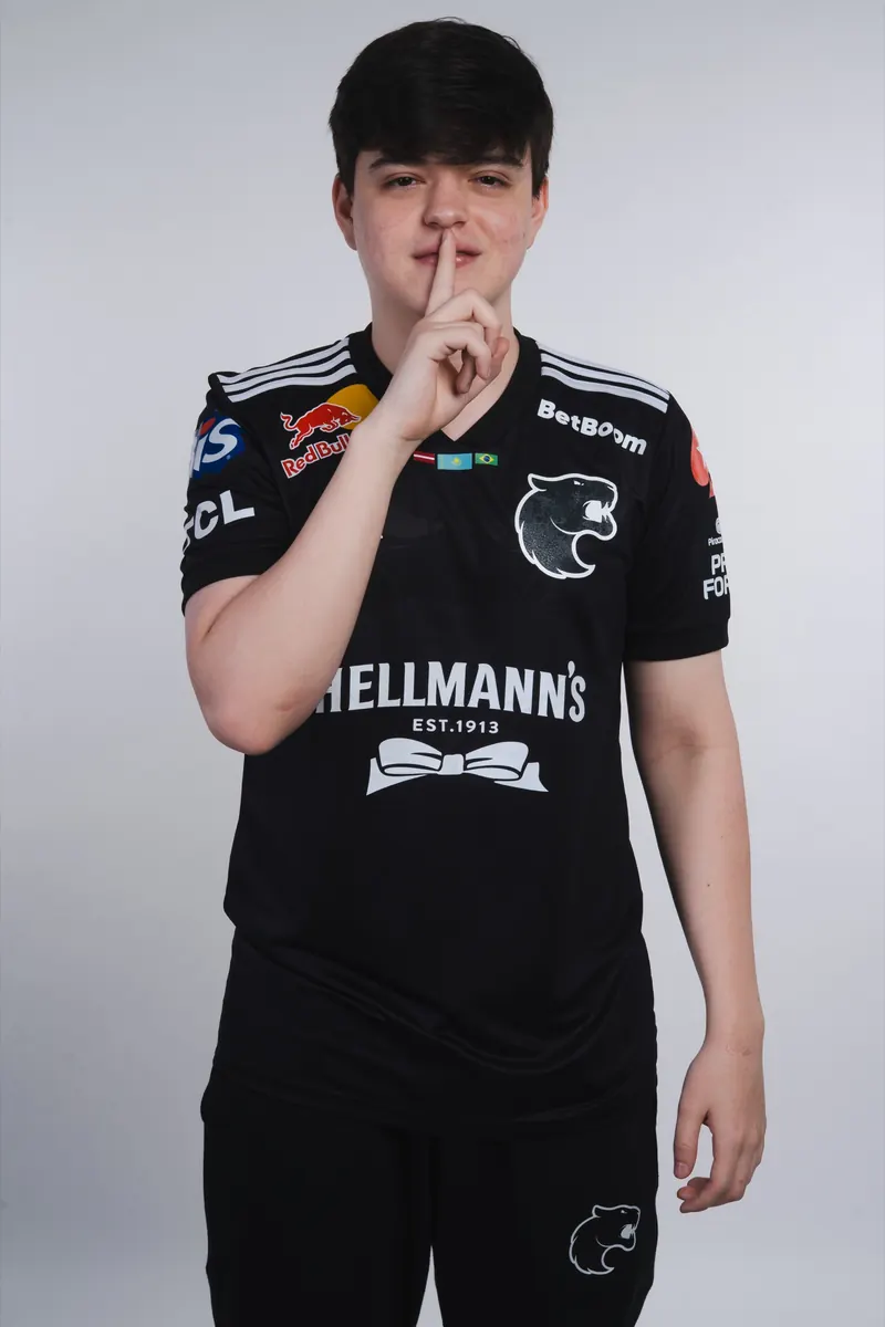 Retrato de jogador de camisa preta da FURIA com o dedo indicador sobre os lábios, em fundo cinza claro.