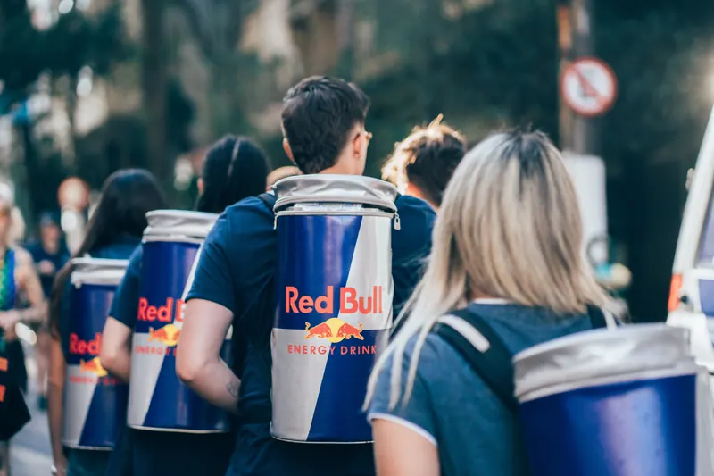 Equipe de costas, com latas gigantes de Red Bull nas mochilas, caminha por uma rua arborizada.