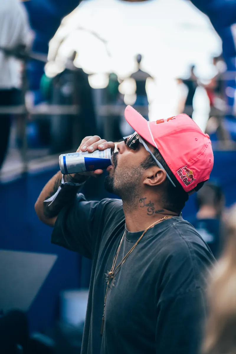 Homem de boné rosa, óculos escuros e camiseta cinza, com uma corrente dourada, bebe de uma lata de Red Bull diante de um palco desfocado em azul.